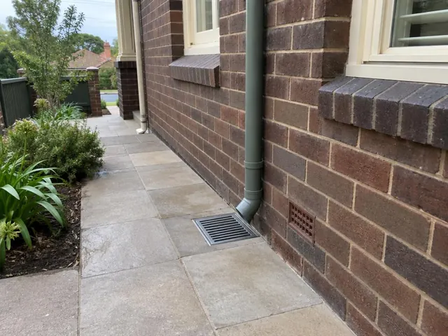 Outdoor drain gully beside a brick house with paved path and garden bed in Fairlight
