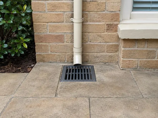 Stormwater grate beside a brick wall with a downpipe over dry paving in Fairlight